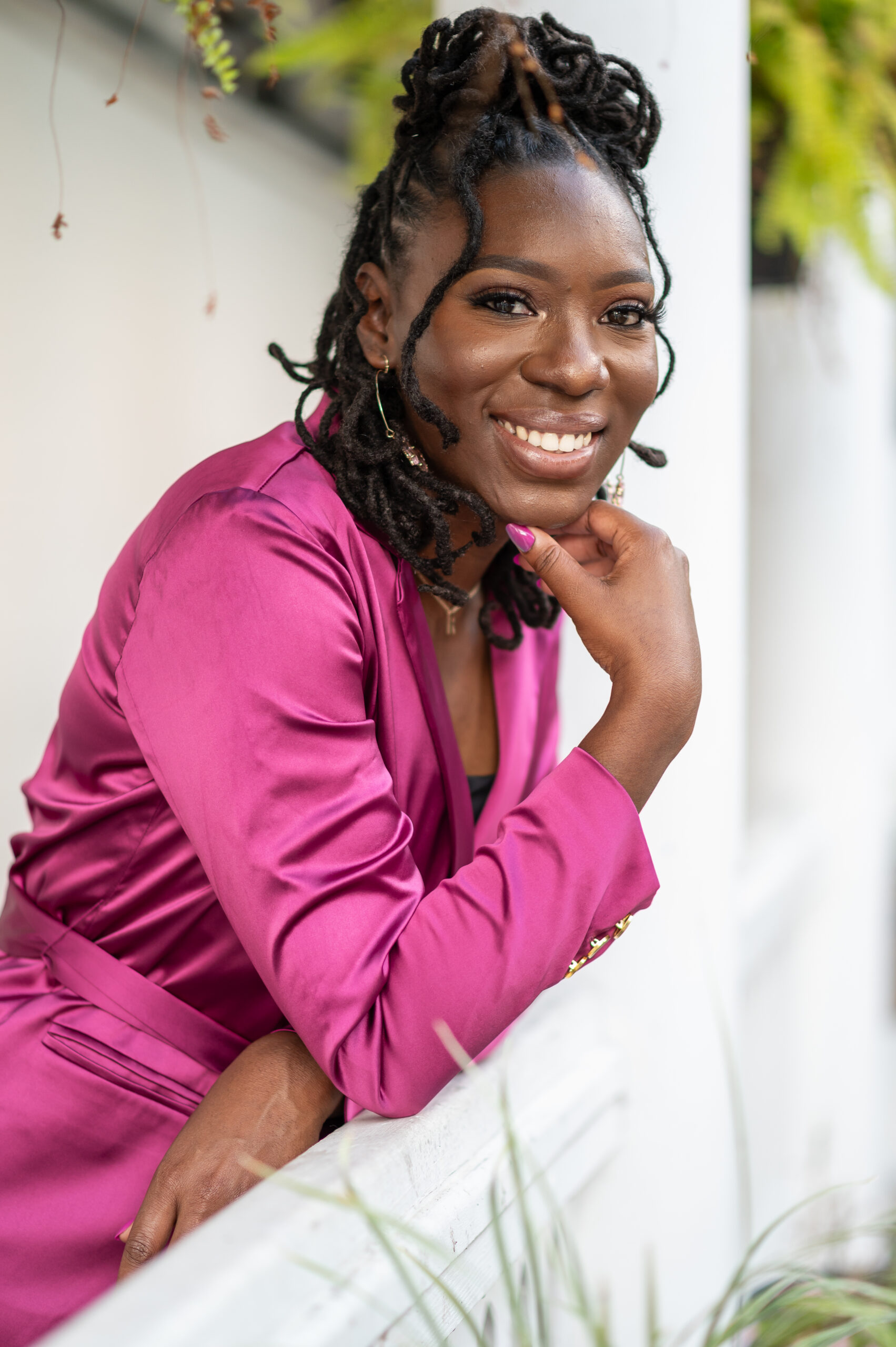 Ronda Taylor, a black woman with her hair in a half-up half-down style, leans over a low wall, rests her chin on her hand and smiles. Ronda wears a red violet silk dress with gold jewelry.