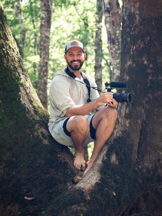 Marsh Dean, a white man with a dark beard wears a ball cap, smiles. He crouches between two trunks of a tree and holds a camera.