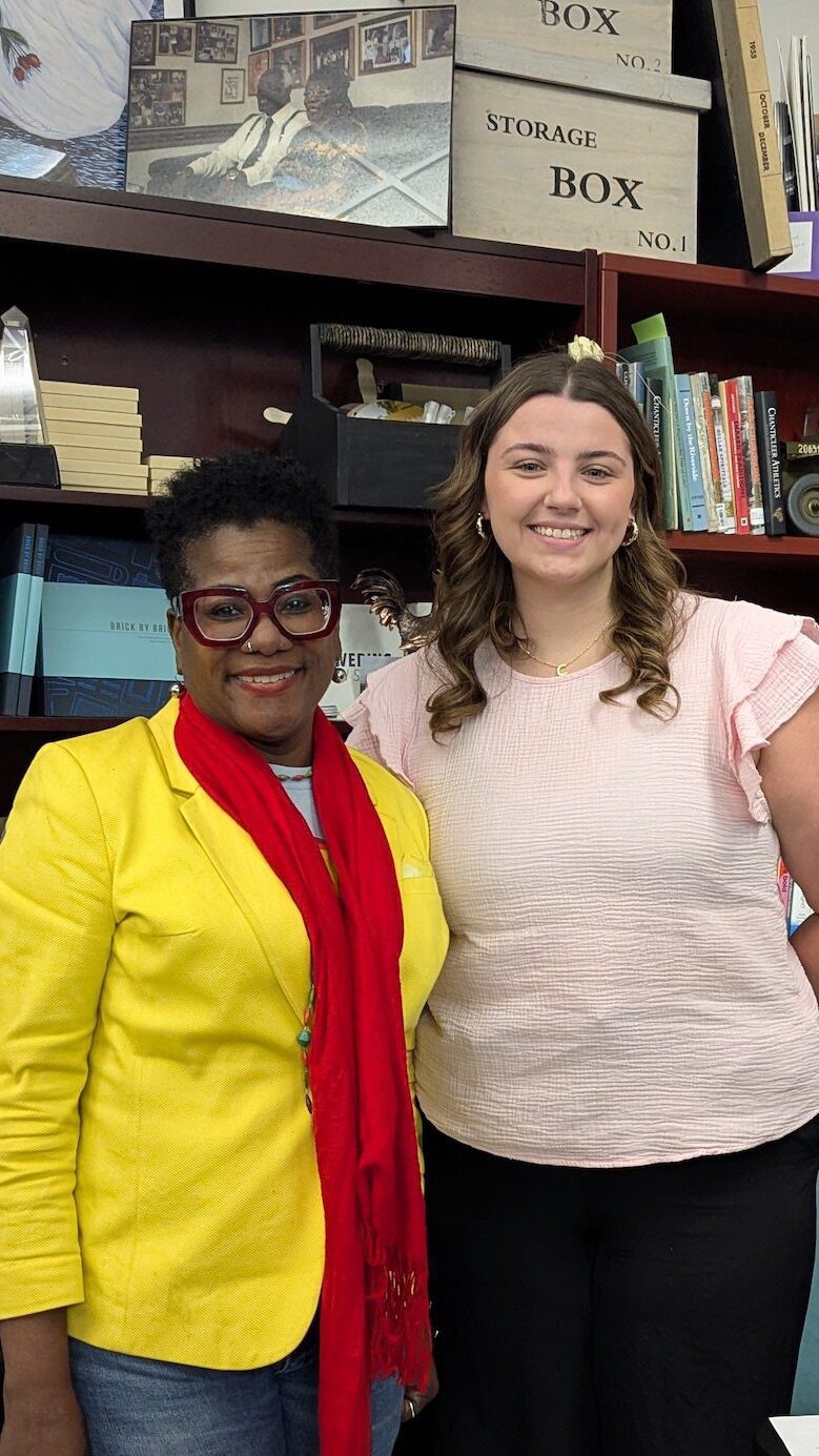 Two women pose in front of a bookshelf in a university office. On the right, a black woman with neon yellow jacket and red scarf with short cropped hair; on the left, a younger white woman with brown curly hair and a pink blouse.